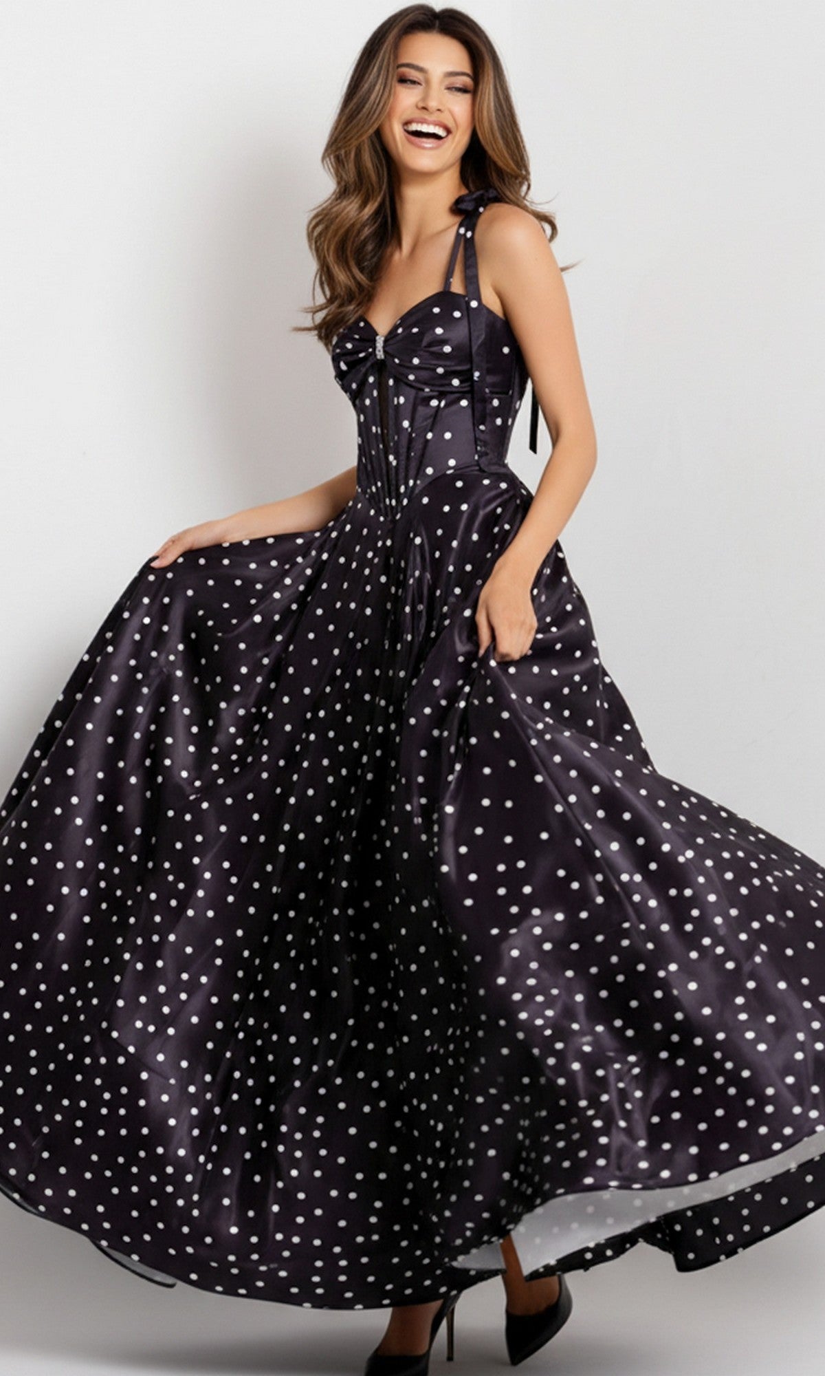 A woman smiles and poses in the Jovani 49774 long black polka-dot satin prom dress, featuring a corset bodice and holding out the skirt.